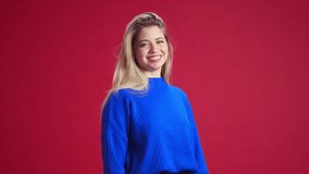 Beautiful young woman with blonde hair, wearing blue sweater, looking at camera and smiling, laughing against red studio background. Happiness. Concept of human emotions, youth, fashion, expression
