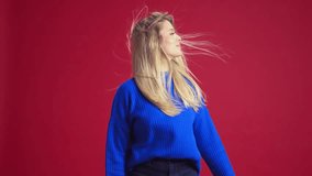 Beautiful, elegant young woman wit blonde hair, posing in blue swear, smiling against red studio background. Wind blowing. Concept of human emotions, youth, fashion, expression