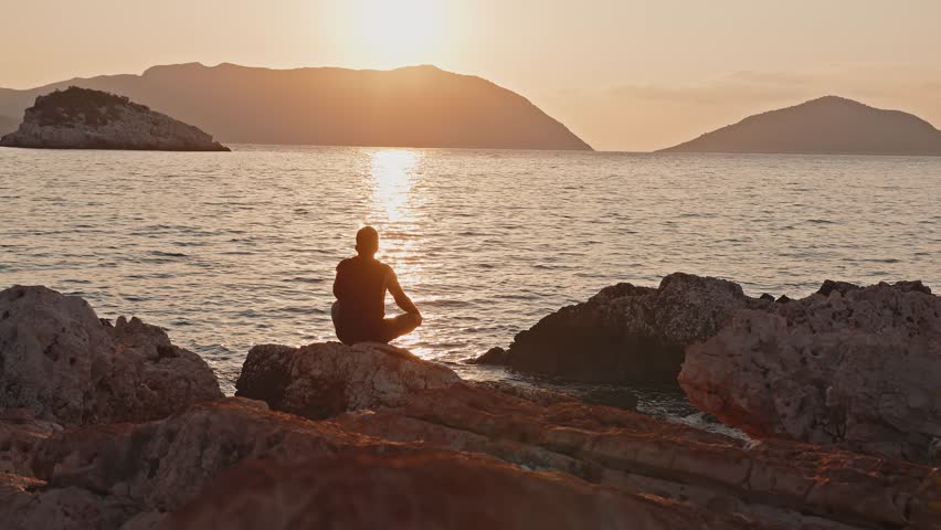 Wide shot. A man meditates at sunrise, sitting on a stone on the seashore.