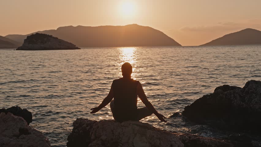 Wide shot. A man meditates at sunrise, sitting on a stone on the seashore.