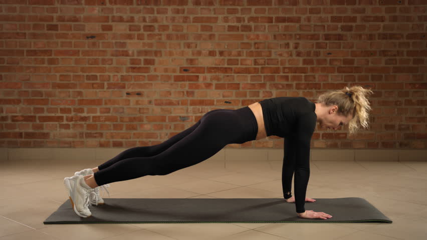 In a minimalist gym, a focused woman in a black outfit expertly performs plank shoulder taps, targeting core and upper body strength. Camera 8K RAW.