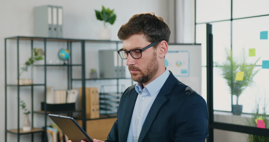 Portrait of attractive positive businessman working on tablet pc and then taking off his glasses and looking at camera in modern office.