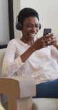 Vertical video of happy african american woman wearing headphones making video call with smartphone. domestic lifestyle, enjoying leisure time at home.
