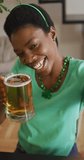 Vertical video of happy african american woman drinking beer making st patricks day video call. domestic lifestyle, enjoying traditional celebration at home.