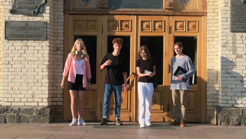 Cheerful students group leaves college building together on sunny day. Friends happy of classes ending walk across campus yard slow motion