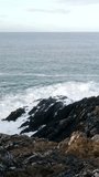 A rocky coastline, where jagged rocks stand firm against the relentless advance of ocean waves. The deep blue sea and the white foam from the waves. Vertical video, pan.