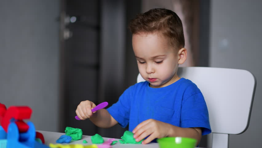 Calm concentrated baby boy uses a stick and a piece of plasticine sitting at desk. Lovely kid plays with dough at home.
