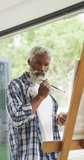 Vertical video of senior african american man painting at home. healthy, active retirement at home.