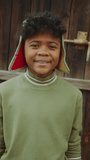 Cheerful African-American boy standing against wooden door in the garden, looking at camera and happily smiling. Video portrait, vertical clip