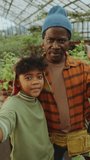 Cheerful African-American grandfather and little boy standing together in a greenhouse farm, waving and talking on camera when video calling or filming vlog. Vertical format shot