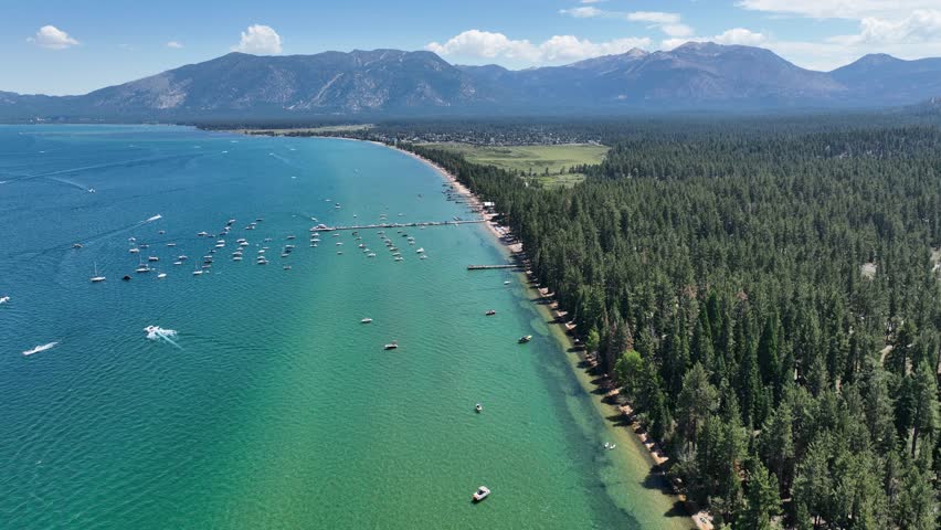 Lovely view of the wonderful blue Lake Tahoe with boats on the shore. A sunny sandy beach and a forest of pine trees on the waterfront. Mountains in the background