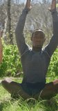 Vertical video of relaxed african american woman practicing yoga sitting in countryside. healthy, active outdoor lifestyle and leisure time.