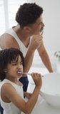 Vertical video of happy biracial father with son brushing teeth at home. spending time at home.