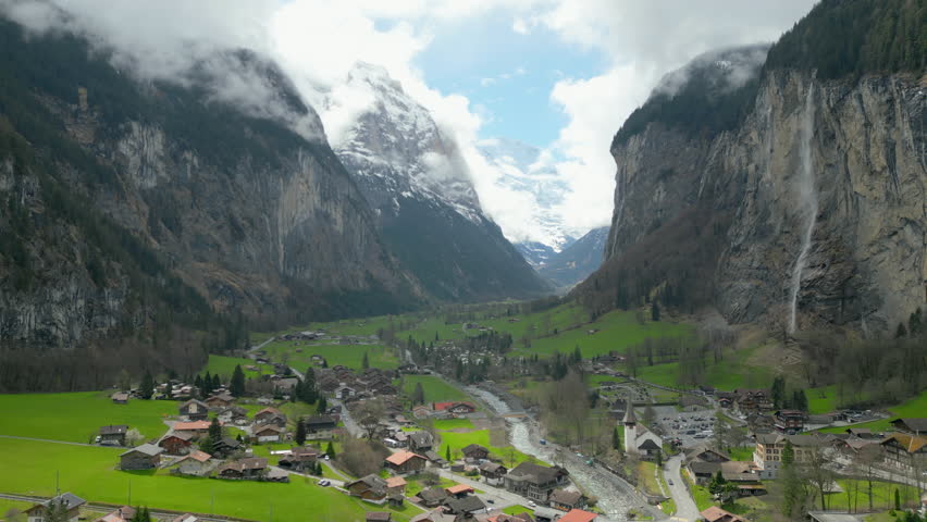 A breathtaking view of a lush Swiss Alpine valley surrounded by steep cliffs, with a cascading waterfall and low-hanging clouds and snowy mountains. Lauterbrunnen, Switzerland, Slow Motion, 4K RAW.  Royalty-Free Stock Footage #3452835283