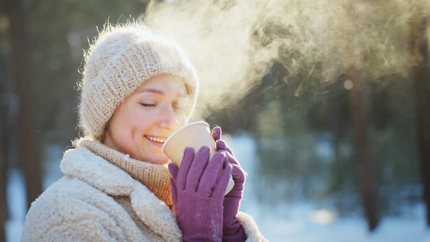 Woman in mitten gloves drinking hot drink in snow covered forest.
