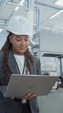 Vertical Portrait of a Black Female Engineer in Hard Hat Walking and Using Laptop Computer at Electronic Manufacturing Factory. Technician Working on Daily Tasks and Research and Development Data.