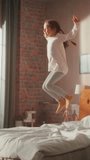 Vertical Screen: Adorable Girl Having Fun at Home, Running Around and Jumping on a Bed in Stylish Bedroom in Loft Apartment. Happy Child Enjoying Childhood and Playing at Home. Slow Motion Footage
