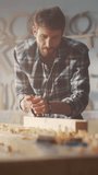 Vertical Screen: Handsome Young Artisan Craftsman in Checkered Shirt Using Hand Plane to Shape a Wood Bar. Carpenter Working on a Project in a Loft Studio with Tools on Walls