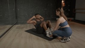 Motivated handsome sporty fit black man with help of lovely female fitness instructor practicing sit-ups exercise, strengthening abdominal and core muscles during indoor sports training. - Powered by Shutterstock - Get 15% off with code: PIKWIZARD15