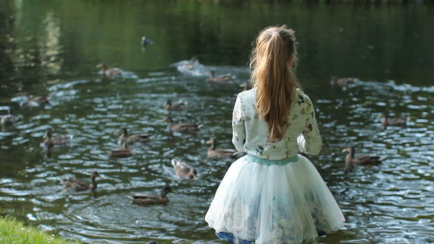 A teenage girl plays on the shore of the lake - feeds the birds, rejoices and rests. Summer park with beautiful lakes, the child walks on the shore of the pond.