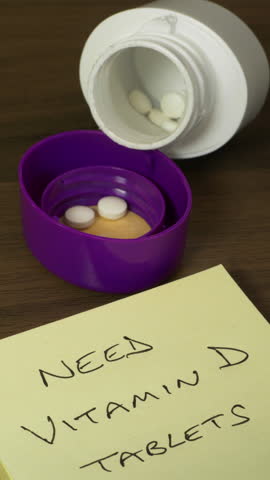 Vertical video – Closeup pan shot of a nearly empty container with a few white tablets or pills of healthcare wellbeing supplement, next to a written note ‘Need Vitamin D tablets’.