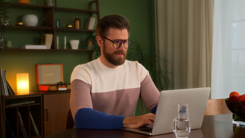 young contented man with beard and eyeglasses sits at table indoors and works on laptop typing on keyboard. pleased ambitious student does online education. remote specialist advanced training. Royalty-Free Stock Footage #3453003771