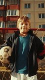 Vertical Shot: Portrait of a Handsome Boy Wearing a Sport Coat Holding a Soccer Ball in the Neighborhood. Young Football Player Looking at Camera, Smiling. Standing in Front of a Old Gate