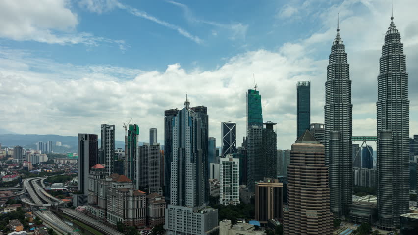 cityscape of Kuala Lumpur featuring the famed Petronas Twin Towers amid a dense cluster of skyscrapers, urban architecture, and lush hills in the backdrop on a cloudy day