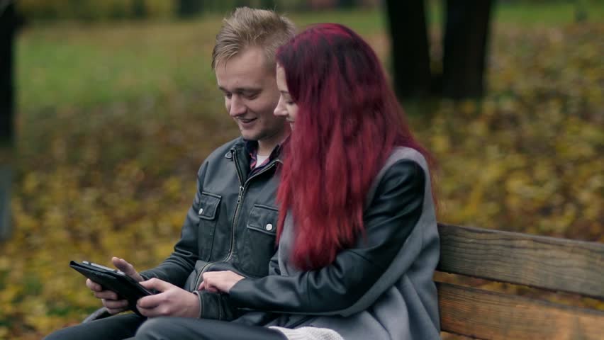 Cute couple sitting on a bench in autumn park and using a digital tablet together. Young woman with red hair and attractive man in a leather jacket discussing someting