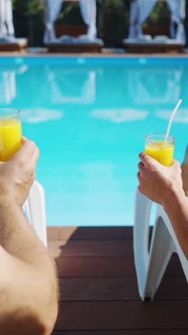 Rear view hands of couple clinking glasses of orange juice when resting on poolside lounge chairs at resort hotel. Man and woman having drinks by the pool. Vacation, relaxation, recreation concept