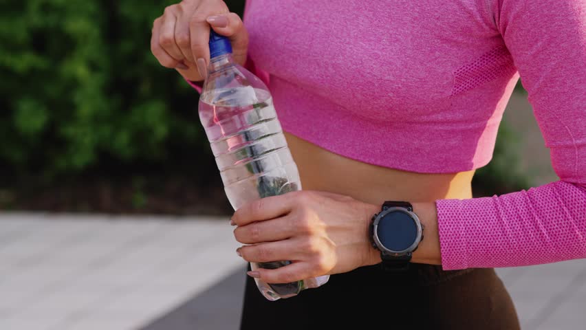 A young sportive woman rests after workout, open a bottle and quenches thirst outdoors in the city park area. A charming and inspired girl athlete with beautiful smile poses for a drinking water