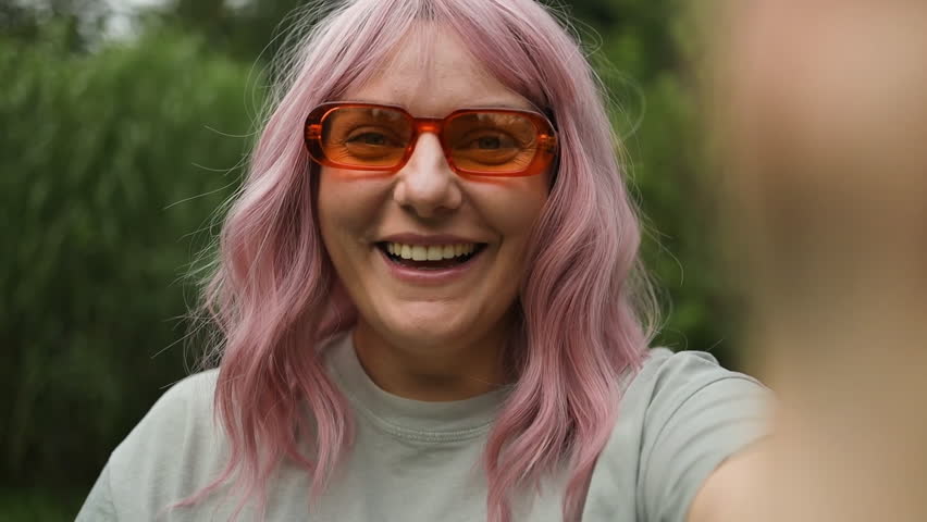 Caucasian joyful smiling beautiful young pink hair woman in orange sunglasses recording video for blog. Female talks on video call through webcam at backyard and waving hand  Royalty-Free Stock Footage #3453265593
