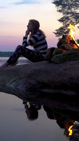Young woman sitting by the bonfire burning in a forest on the shore of a lake at midnight on the Summer Solstice. Summer White Night in Finland. Sound of campfire and wildlife, birdsong Royalty-Free Stock Footage #3453298847