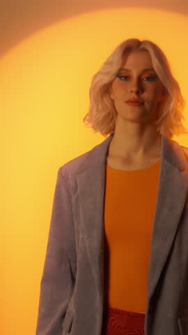 Blonde elegant woman in jacket walking and posing arms crossed over orange colour wall in studio