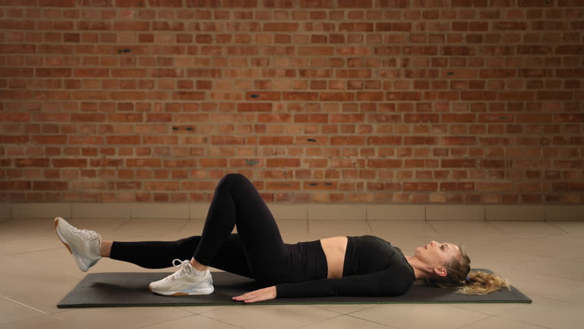 An expert fitness coach meticulously executes the right single-leg hip bridge, focusing on asymmetrical strength development and muscular balance against a rustic brick wall in a modern training space Royalty-Free Stock Footage #3453361621