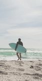 Vertical video of biracial man alone with surfboard on sunny beach. healthy, active retirement beach holiday.
