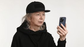 A senior woman shows signs of disappointment while using her smartphone for a video call, dressed in a black hat and turtleneck, with a minimalist white background. 