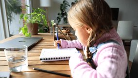 A smart little Caucasian girl sitting at the table at home paints in a notebook near the window. Learning, education concept.
