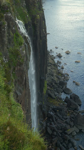 VERTICAL: Beautiful big waterfall falling from a grassy cliff onto a rocky shore covered with granite stones. Breathtaking scenery with rugged ocean coastline near famous Neist Point on Isle of Skye.