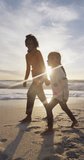 Vertical video of happy biracial mother and daughter walking on sunny beach. healthy, active time beach holiday.