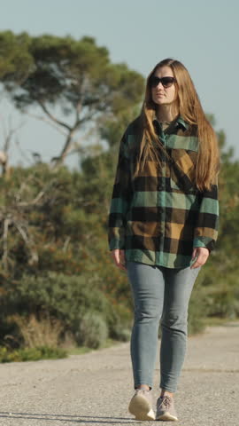 Vertical video. A young woman walks along an abandoned highway through the forest, walking in front of her.