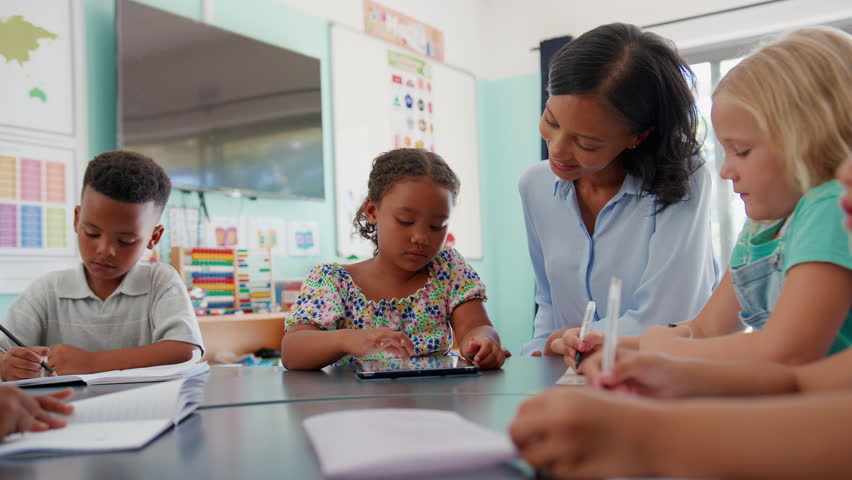 Female primary or elementary school teacher helping students with digital tablet around table in class - shot in slow motion - Powered by Shutterstock - Get 15% off with code: PIKWIZARD15