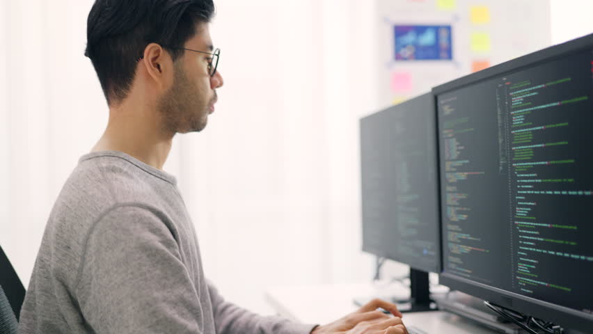 Side profile of an Asian male software developer working attentively on complex code across triple monitors in a bright office. Royalty-Free Stock Footage #3453549679