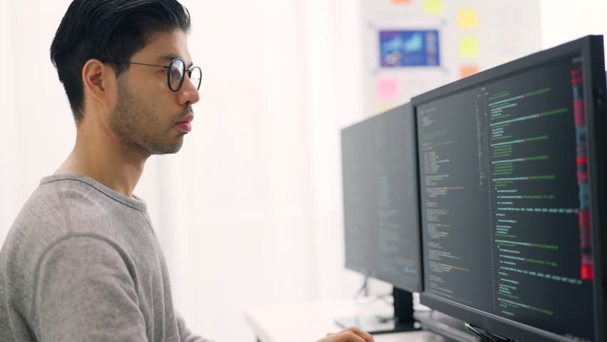 Side profile of an Asian male software developer working attentively on complex code across triple monitors in a bright office.