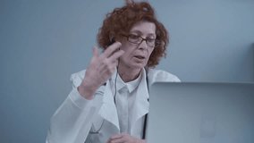 Female senior doctor therapist in white coat and headset talking video conferencing on laptop in office using online video calling application. Remote medical care for patients, online consultation.