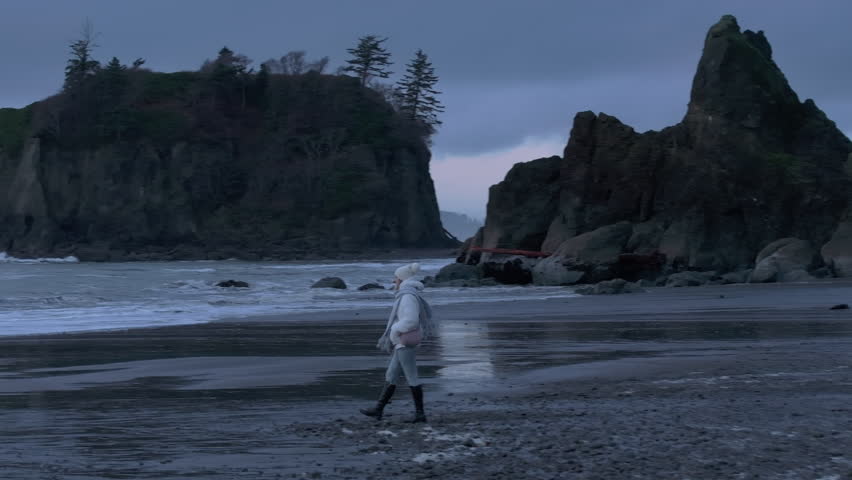 Dramatic nature aerial 4k. Female tourist exploring Washington coast on gloomy grey day. Scenic black sea rocks at Pacific ocean coast on overcast evening. Woman contemplating at cinematic Ruby beach