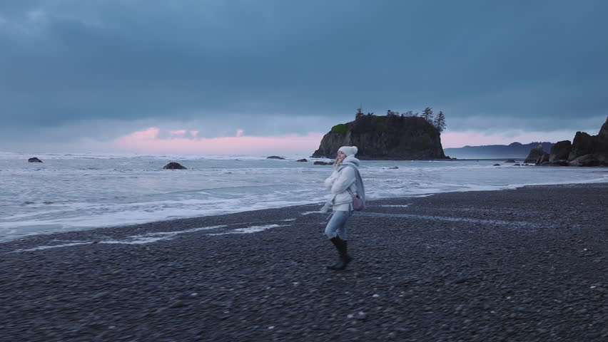 Female tourist exploring Washington coast on gloomy grey day. Scenic black sea rocks at Pacific ocean coast on overcast evening. Woman contemplating at cinematic Ruby beach. Dramatic nature aerial 4k