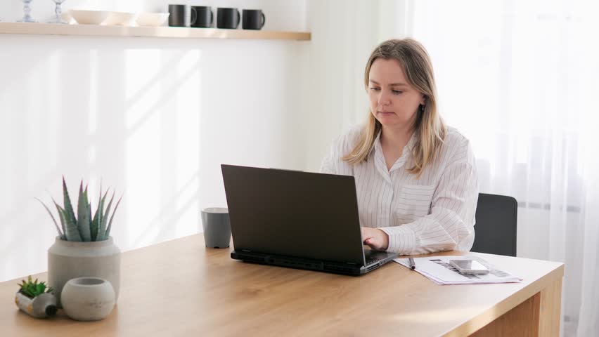 Female journalist sits bright living room with laptop, finishing morning coffee. Journalist types on laptop, periodically taking sip aromatic drink. Laptop becomes working tool comfort of home office. Royalty-Free Stock Footage #3453623475