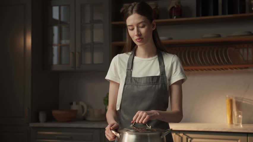 A young woman dressed in an apron stands in a home kitchen, savoring the scent of a dish she is preparing. The gentle evening light filters in through the window, highlighting the steam rising from
