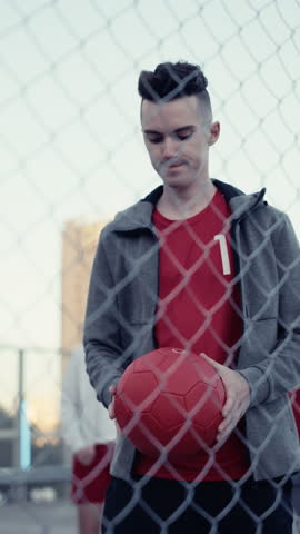 Vertical Screen: Portrait of a Handsome Young Football Player Looking at Camera, Spinning a Red Soccer Ball on His Finger. Stylish Footballer Standing Behind a Fence with a Group of Diverse People Royalty-Free Stock Footage #3453652739